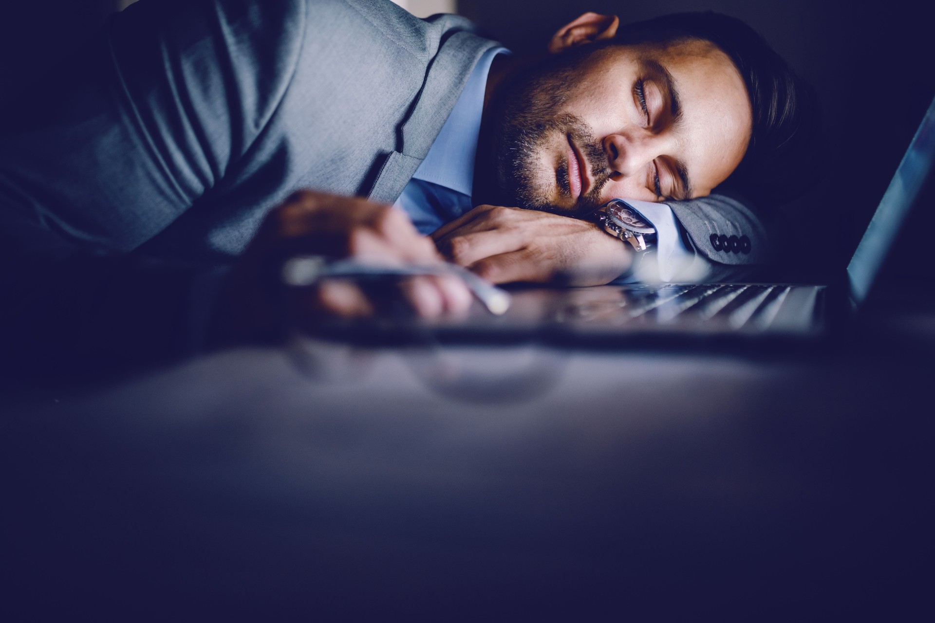 Exhausted Caucasian businessman in suit sleeping on desk in office. On desk is laptop. Overworking concept.